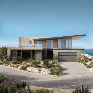 A modern house with large windows and a balcony, surrounded by desert plants and rocks, near a coastline.
