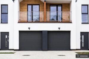 A modern building facade featuring two garage doors and upper balcony with glass railing.