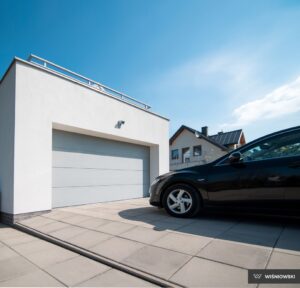 A modern garage with a light gray door, next to a black car parked on tiled pavement under a blue sky.