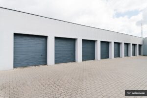 A row of six blue garage doors on a light wall with a gray cobblestone floor.