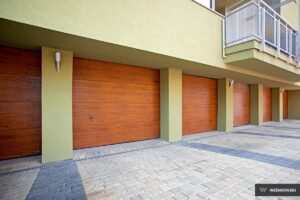 Three wooden garage doors on a green wall, with a stone pavement below.