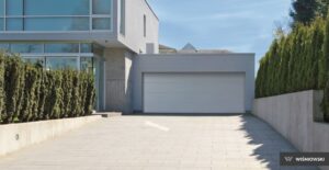 The image features a modern, sleek garage door with a full-view design, characterized by large glass panels, situated in a residential environment. The garage door appears to be stylishly integrated into a contemporary home's exterior, offering a seamless blend of functionality and aesthetic appeal. The full-view glass panels are framed with a metallic finish, enhancing the overall modern look while allowing natural light to illuminate the garage space.
